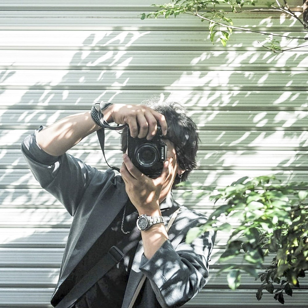 Portrait du photographe contemporain Yusuke F tenant un appareil photo devant un rideau métallique ombragé par des feuillages.