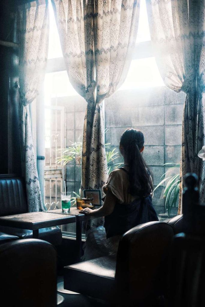 Femme assise seule dans un café, observée de dos près d’une fenêtre lumineuse.