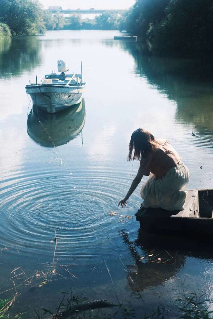 Femme accroupie au bord d’une rivière, la main dans l’eau, avec une barque en arrière-plan.