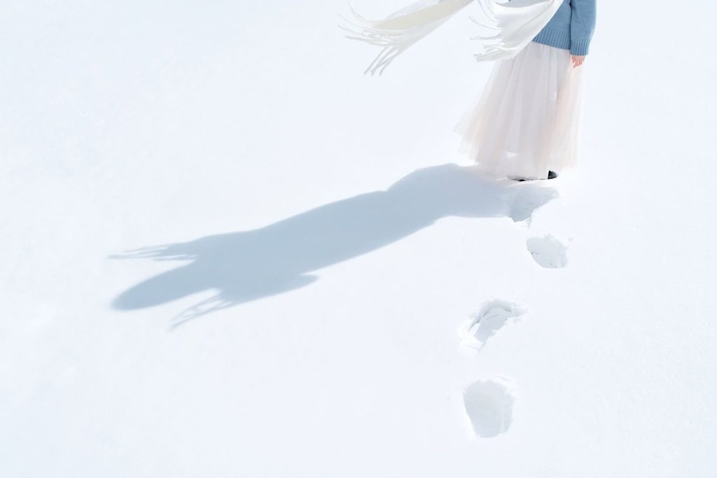 Personne vêtue de blanc marchant dans la neige, laissant des empreintes et une longue ombre.