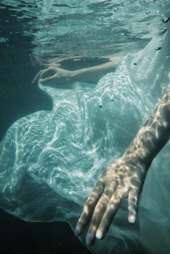 Femme en robe blanche immergée sous l’eau, le bras tendu vers la surface.