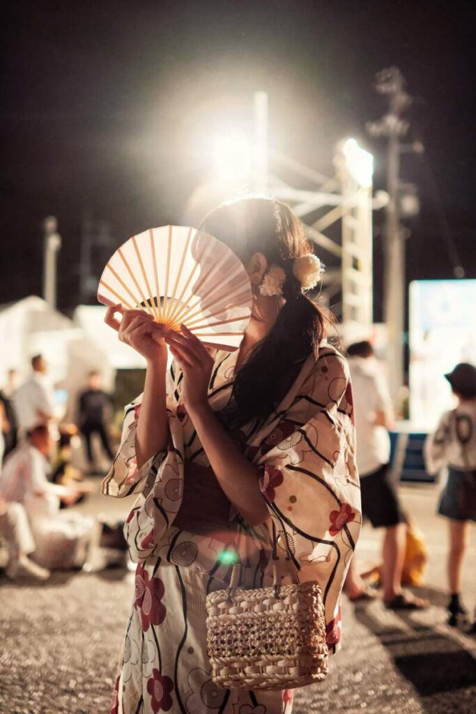 Femme en yukata tenant un éventail lors d’une fête nocturne, éclairée à contre-jour.
