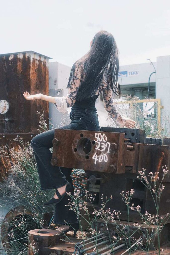 Jeune femme assise sur une structure métallique rouillée dans un paysage urbain, les bras ouverts.